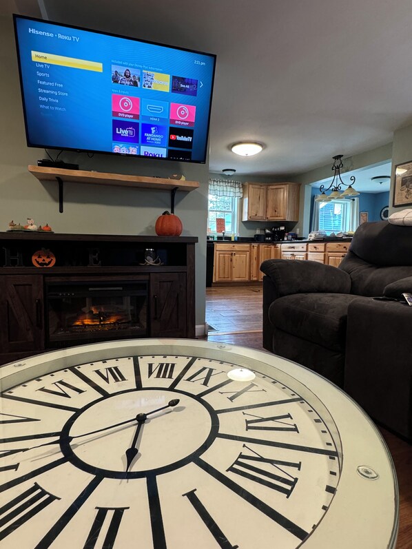A living room with a large clock, a fireplace, and a television displaying a Roku TV interface. - Northern Maine Adventure (Fort Fairfield)