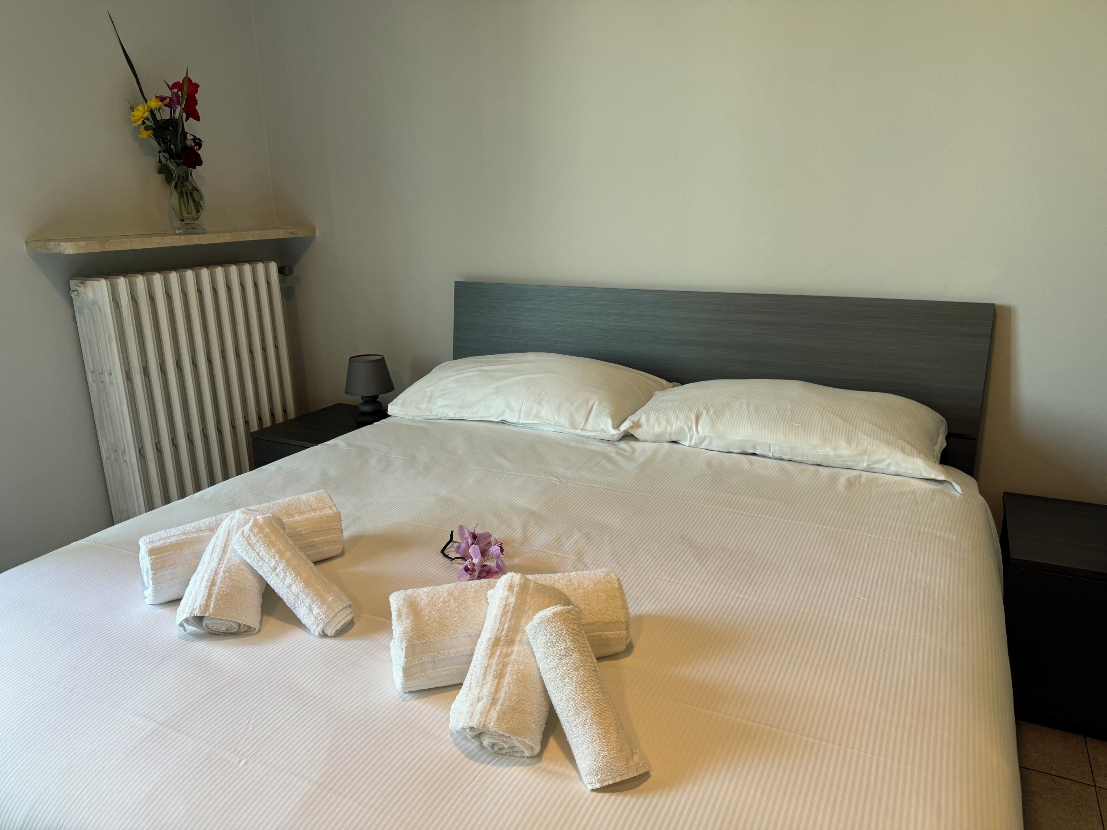 A neatly made bed with white linens and towels arranged in a heart shape, a bedside lamp, and a vase with flowers on a nightstand.