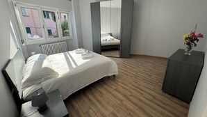 A modern bedroom with a large bed, a bedside table with a lamp, a mirror, and a small side table with a vase of flowers.