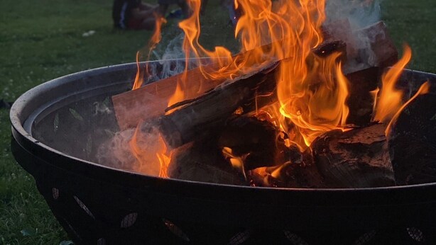 Una fogata con leños ardiendo, rodeada de un área verde con un cuerpo de agua y barcos al fondo.