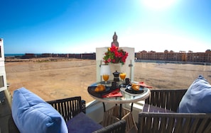 A balcony with a table set for breakfast, overlooking a sandy beach and buildings in the distance. - Sea View Suite Sleeps 2 With Parking (Hurghada)