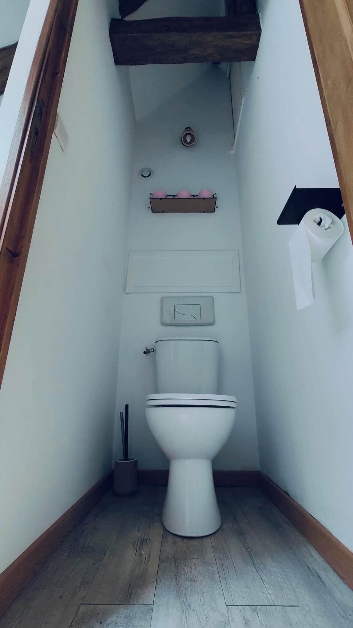 Une petite salle de bain étroite, avec des toilettes blanches, une étagère en bois au-dessus des toilettes et une petite poubelle.