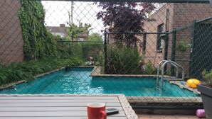 A swimming pool surrounded by a chain-link fence, with a red cup and a remote control on a wicker table in the foreground. - Right in the center across from the stores  (Udenhout)