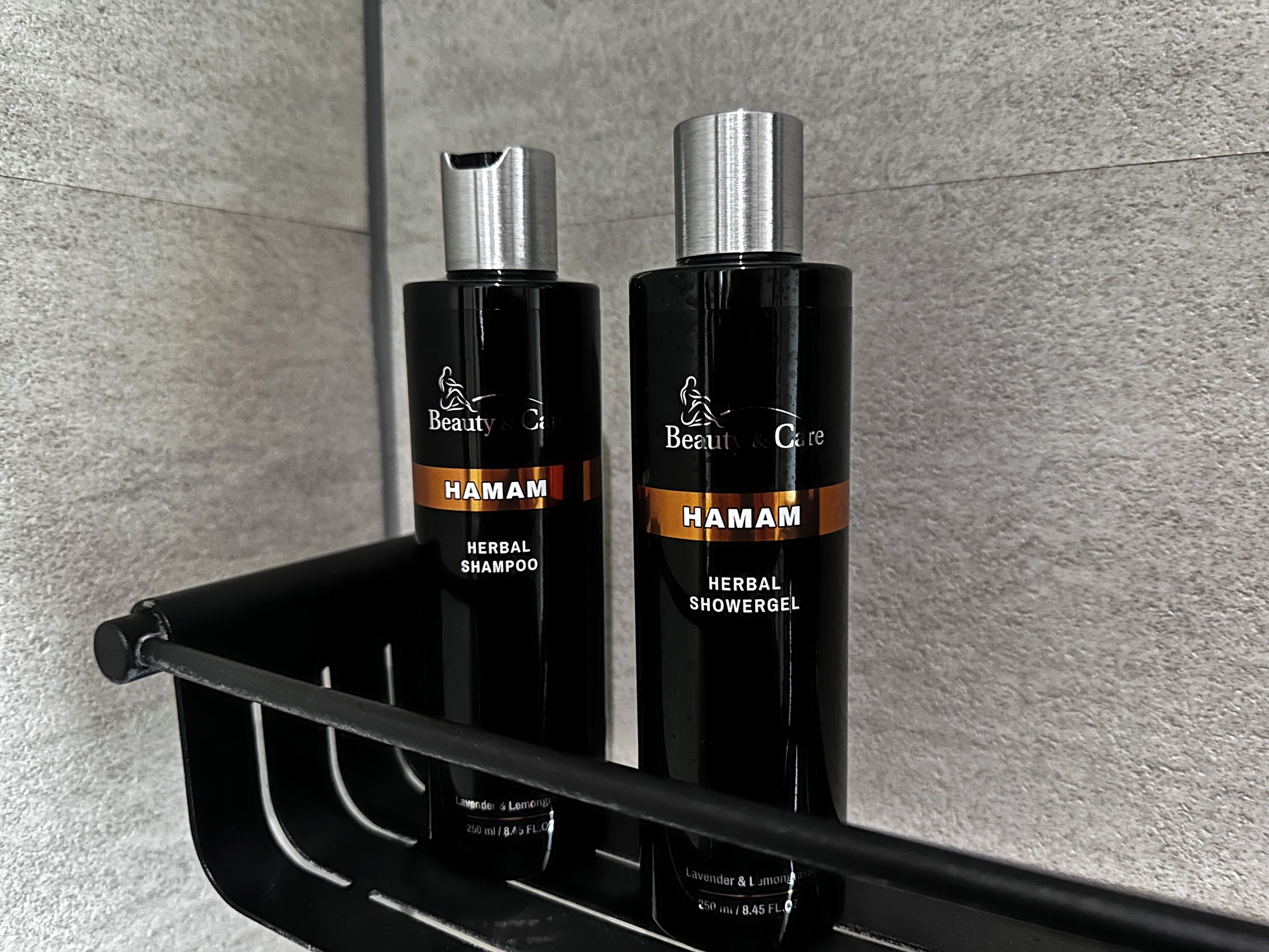 Dos botellas de Champú y Gel de Ducha Herbal Hamam sobre un estante negro frente a una pared con textura.