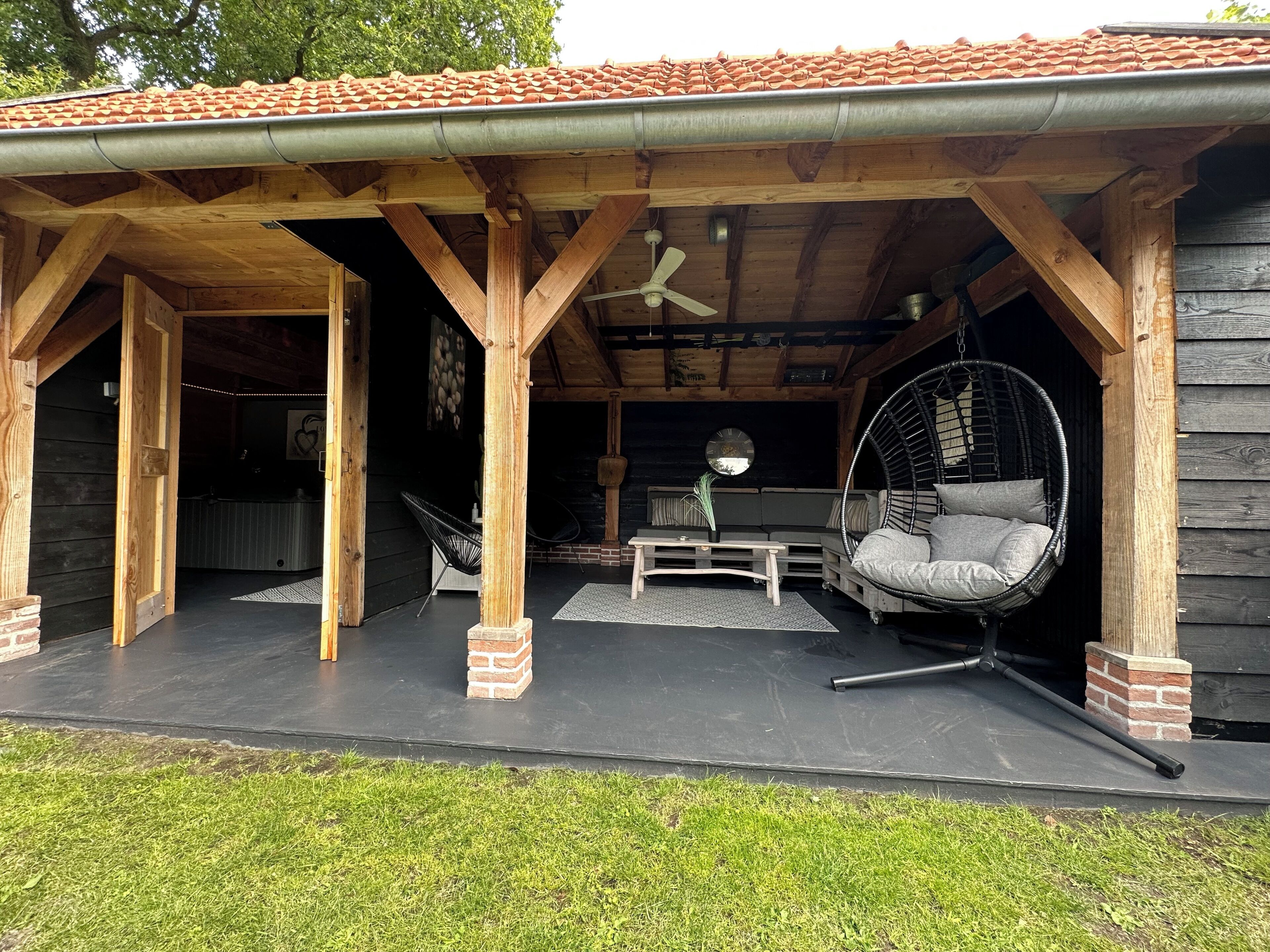 Un espacio al aire libre cubierto con estructura de madera, una silla colgante, una mesa y un ventilador de techo.