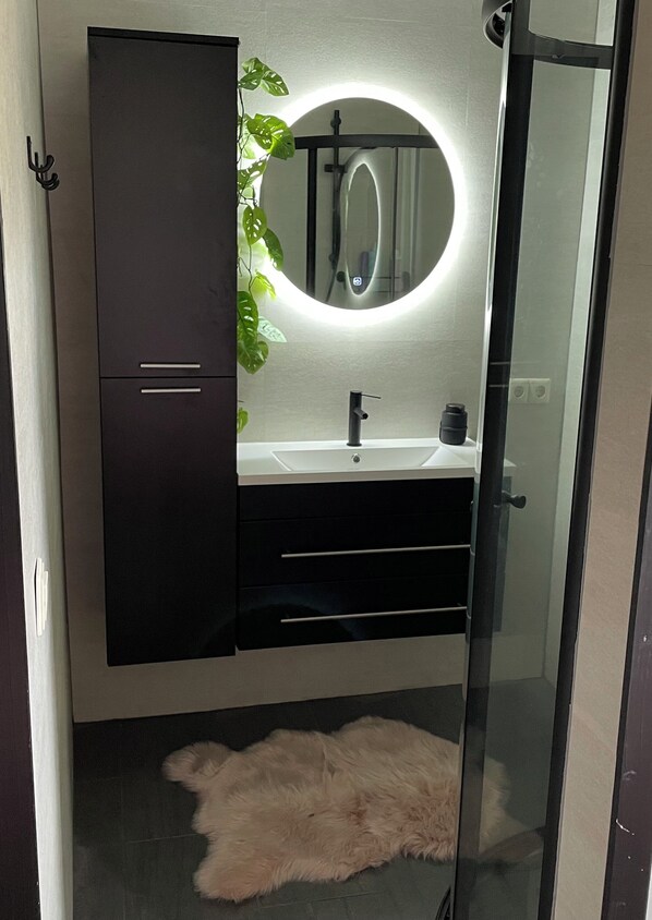 A modern bathroom with a dark cabinet, a round mirror with a light, a sink, and a fluffy rug on the floor.