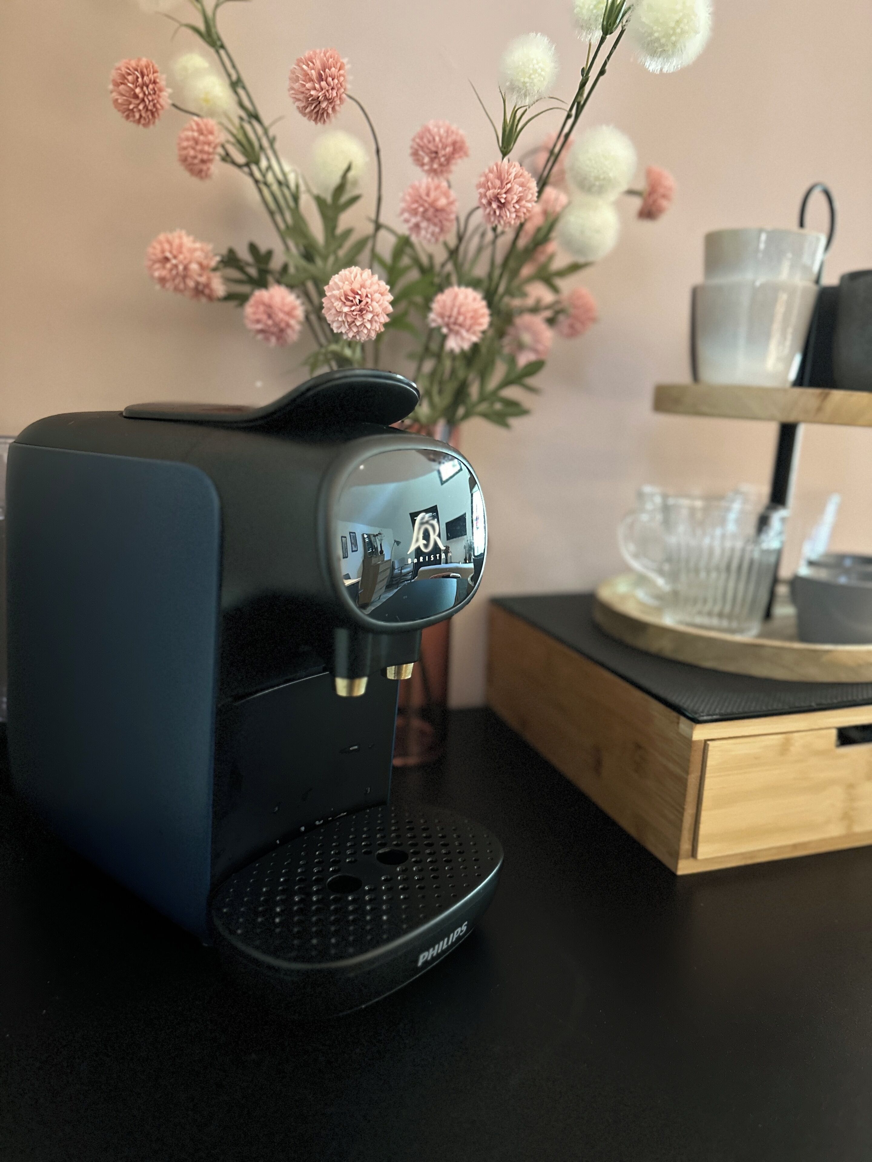 Een zwarte Philips koffiezetapparaat op een zwart aanrecht, met een houten bijzettafeltje en een vaas met roze en witte bloemen op de achtergrond.