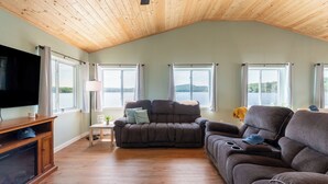 A living room with a wooden ceiling, a television, a sofa, and large windows overlooking a lake. - The Lake Life by Avantstay Lakefront + View + Hot Tub (Pocono Lake)