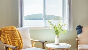 A room with a view of a lake, a white chair, a yellow throw, and a small table with a plant. - The Lake Life by Avantstay Lakefront + View + Hot Tub (Pocono Lake)