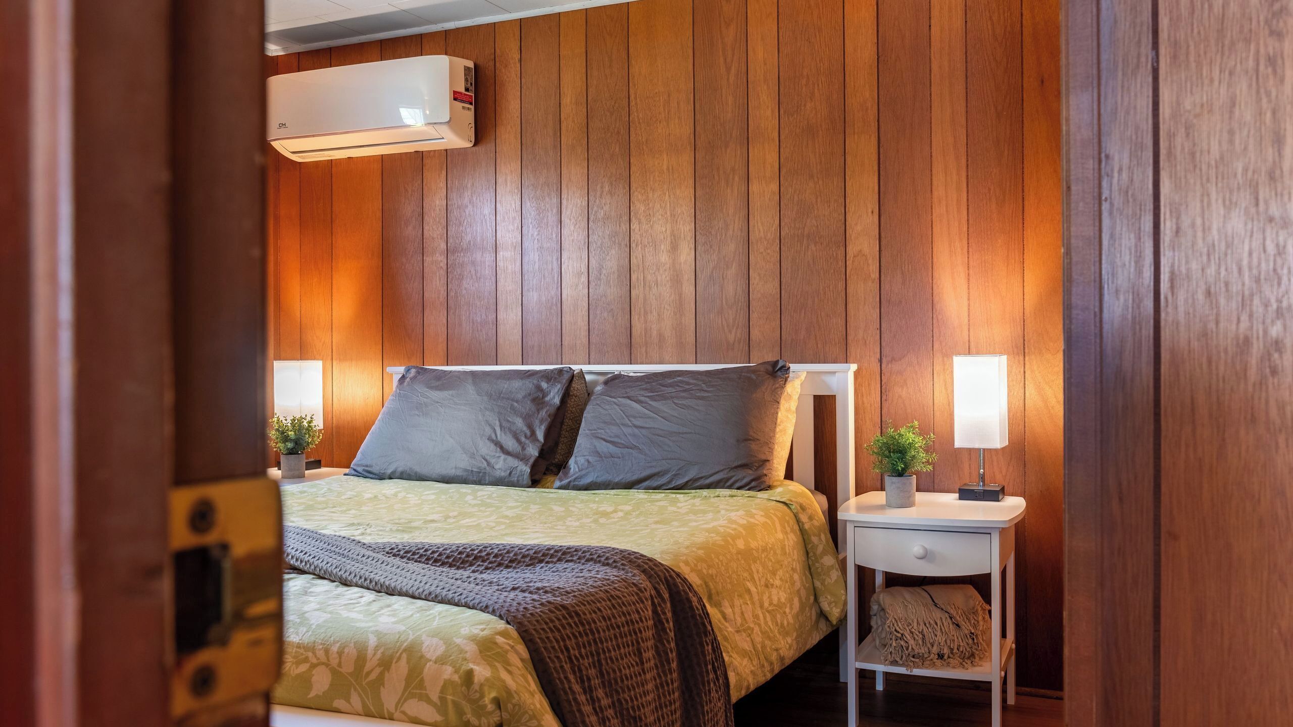 A bedroom with a bed, bedside tables, a nightstand, a wall-mounted air conditioner, and a potted plant.
