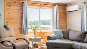 A cozy room with a wooden wall, a window with curtains, a sofa, a chair, a small table with a plant, and an air conditioning unit. - The Lake Life by Avantstay Lakefront + View + Hot Tub (Pocono Lake)