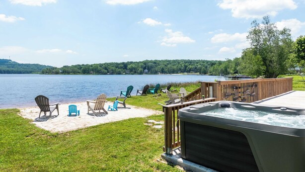 A lakeside patio with Adirondack chairs, a hot tub, and a wooden deck overlooking the water. - The Lake Life by Avantstay Lakefront + View + Hot Tub (Pocono Lake)