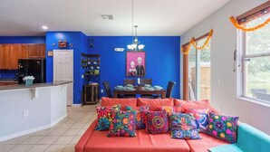 A kitchen with a dining area, a sofa with colorful pillows, and a window with orange decorations.