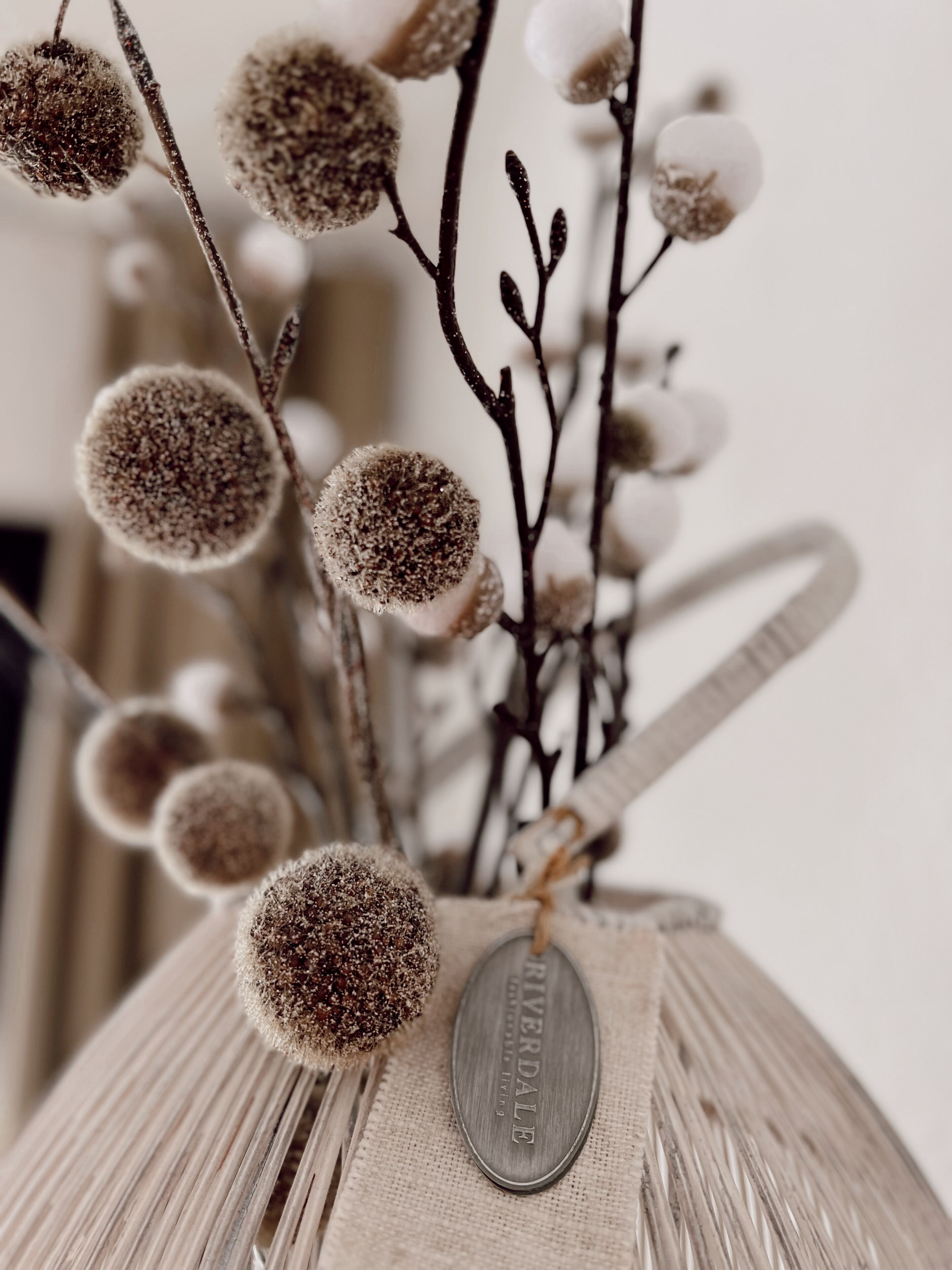 Un florero con base de arpillera y una etiqueta metálica, que contiene ramas con yemas redondas y heladas, y elementos similares al algodón blanco.