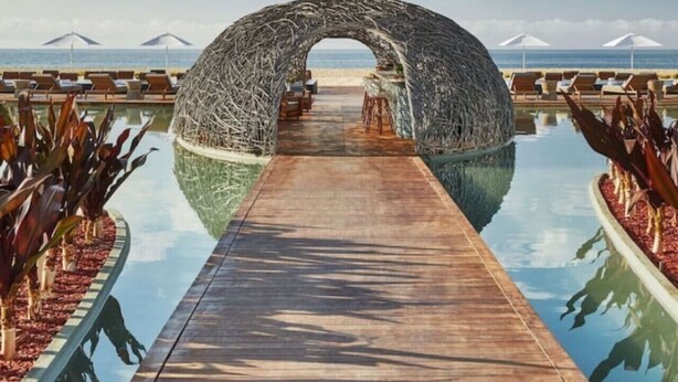 Een houten pad dat leidt naar een strandlounge met een bijzondere, gebogen constructie.