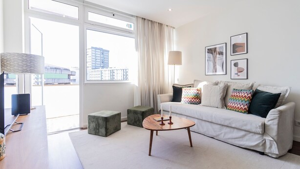 A modern living room with a white sofa, colorful pillows, a wooden coffee table, and a lamp. There is a window with curtains, a small side table, and a framed picture on the wall. - 2BR Wonder in the City Center by LovelyStay (Porto)