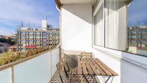 Ein Balkon mit einer Holzbank und einem Lüftungsgitter, mit Blick auf eine Stadtlandschaft mit Gebäuden und einem roten Bus.