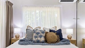 A hotel room with a bed, two bedside tables with lamps, a window with sheer curtains, and an air conditioning unit.