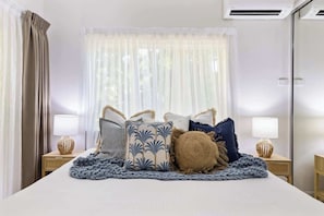 A hotel room with a bed, two bedside tables with lamps, a window with sheer curtains, and an air conditioning unit.