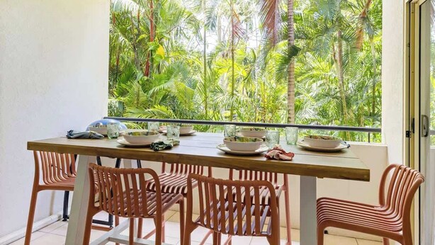A balcony with a view of dense tropical foliage, a wooden table set with plates and glasses, and wicker chairs.