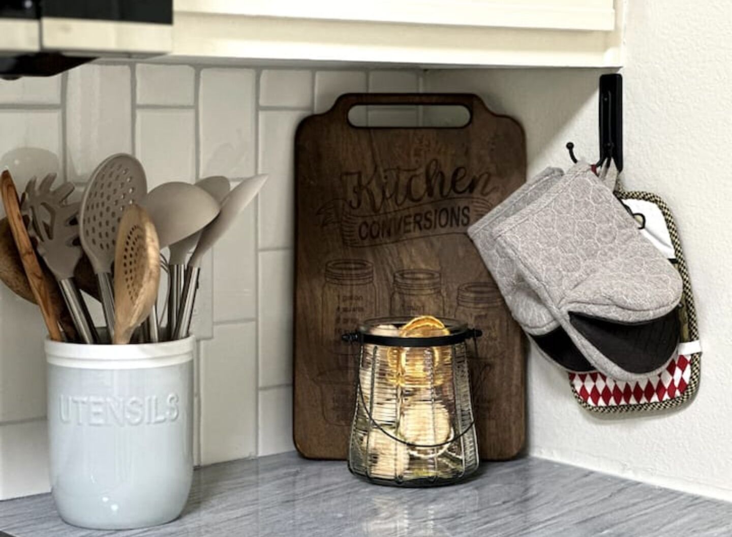 Un piano di lavoro in cucina con un portauova bianco, un tagliere di legno con la scritta 'Kitchen Conversions', un barattolo di vetro con coperchio e una presina decorata appesa al muro.