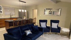 En moderne stue med en mørkeblå hjørnesofa, to ens matchende lænestole og en lille, hvid skammel. En køkkenafdeling med spiseplads kan ses i baggrunden.
