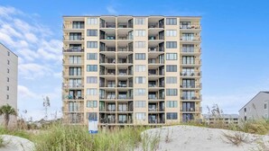 A multi-story residential building with balconies, located near a sandy area with sparse vegetation.