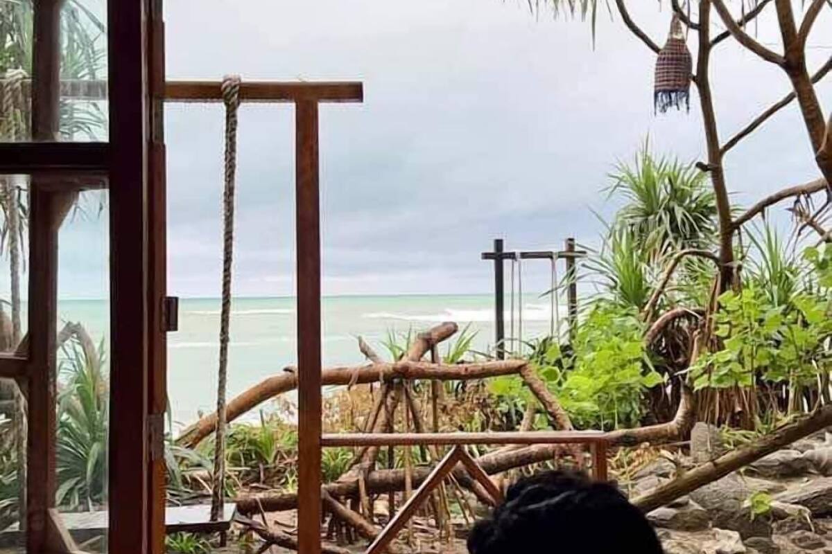 A room with a large window overlooking a beach with wooden structures and plants.