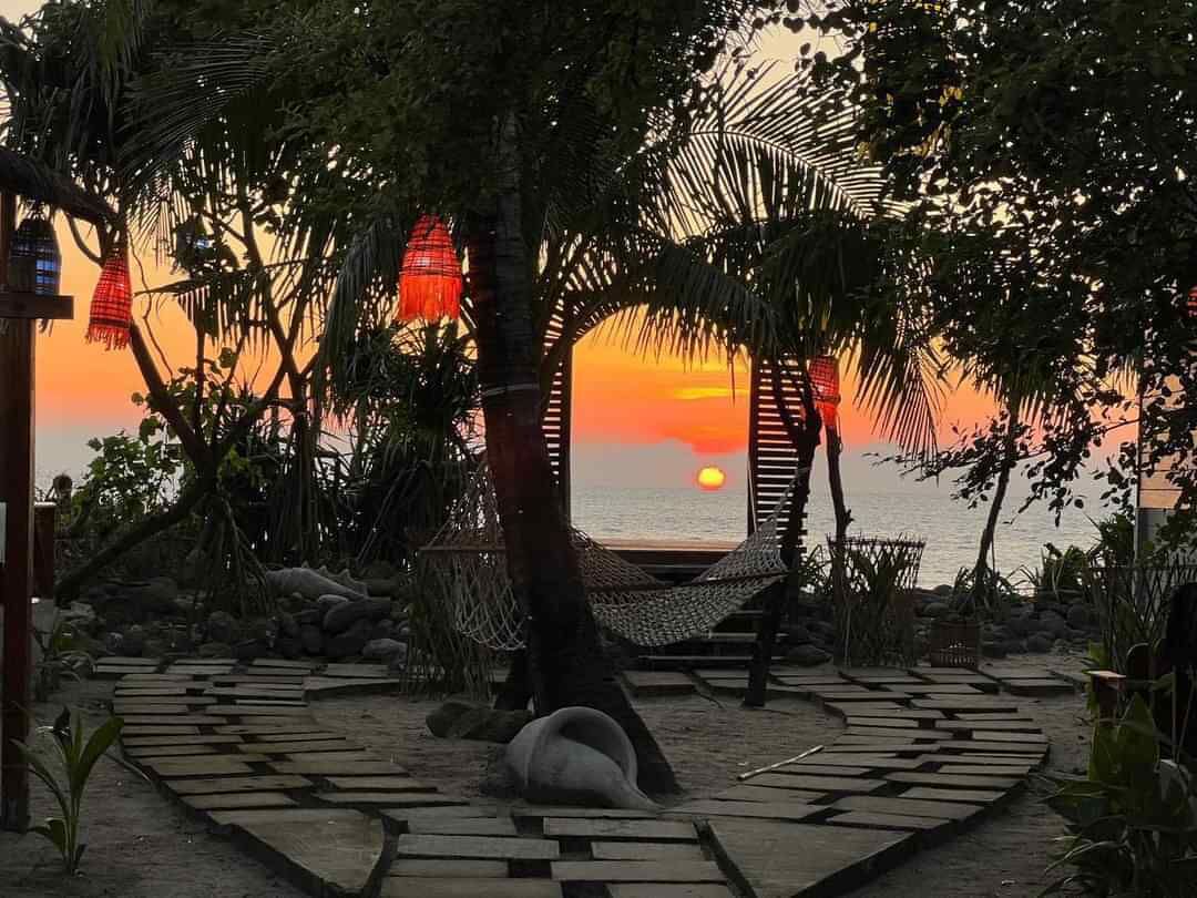 Un hamac sous un arbre, un sentier et un coucher de soleil sur l’eau.