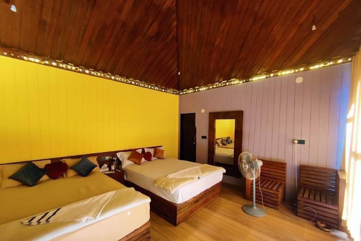 A room with two beds, wooden flooring, a mirror, and a standing fan.
