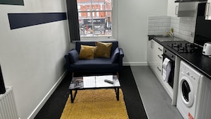 A compact living space with a sofa, a coffee table, a washing machine, and a window with a view of buildings. - London Road Studio Retreat in Leicester (Leicester)