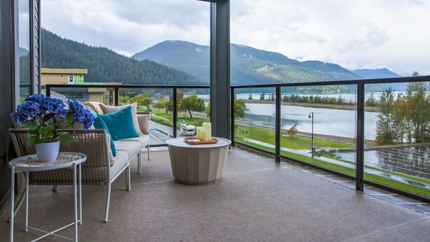 A balcony with a sofa, a table with a potted plant, and a view of a lake and mountains. - The Luna | New Penthouse w Patio, Lake & Mtn Views (Harrison Hot Springs)