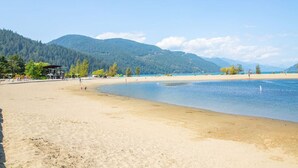 A beach with a clear view of the water, sandy shore, and a forested mountain in the background. - The Luna | New Penthouse w Patio, Lake & Mtn Views (Harrison Hot Springs)