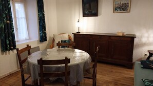 Un comedor con una mesa redonda cubierta con una lona protectora, rodeada de sillas de madera. Un mueble de madera con cajones está apoyado contra la pared, y encima hay un cuadro colgado.