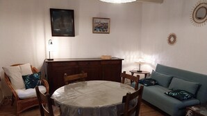 Comedor con una mesa redonda cubierta de plástico, rodeada de sillas de madera. Se observa un sofá y una pequeña mesa con una lámpara al fondo.