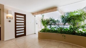 L’entrée d’un hôtel au design moderne, avec une porte numérotée, un sol carrelé et un bac à fleurs contenant des plantes tropicales.