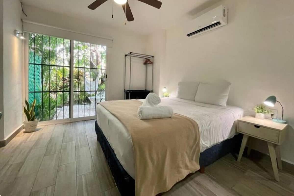 Une chambre à coucher avec un lit, un ventilateur de plafond, une table de chevet, une plante en pot et une vue sur la verdure par la fenêtre.