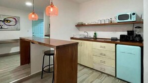 Une cuisine avec un îlot central en bois, un réfrigérateur bleu clair, un four à micro-ondes et une machine à café. Des lampes suspendues orange et un tableau ornent le mur.