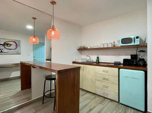 Une cuisine avec un îlot central en bois, un réfrigérateur bleu clair, un four à micro-ondes et une machine à café. Des lampes suspendues orange et un tableau ornent le mur.