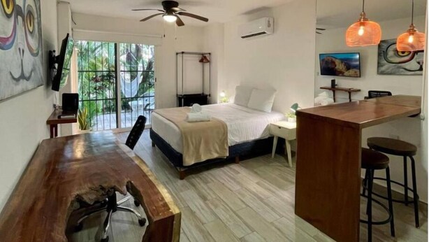 Une chambre moderne avec un lit, un bureau et un coin repas. On y trouve un ventilateur de plafond, un téléviseur et des œuvres d’art accrochées aux murs.