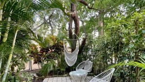 Une terrasse agrémentée d’un fauteuil suspendu et de deux chaises en métal, entourée d’une végétation luxuriante et dotée d’un sol carrelé.