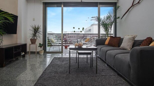 Uma sala de estar moderna com um sofá cinza, uma mesa de centro e um grande painel de vidro deslizante que dá para uma varanda com plantas e vista da cidade.