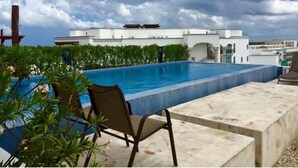 Piscina no terraço com uma cadeira, rodeada por vegetação e um edifício branco ao fundo.