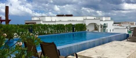 A rooftop pool with a chair, surrounded by greenery and a white building in the background.