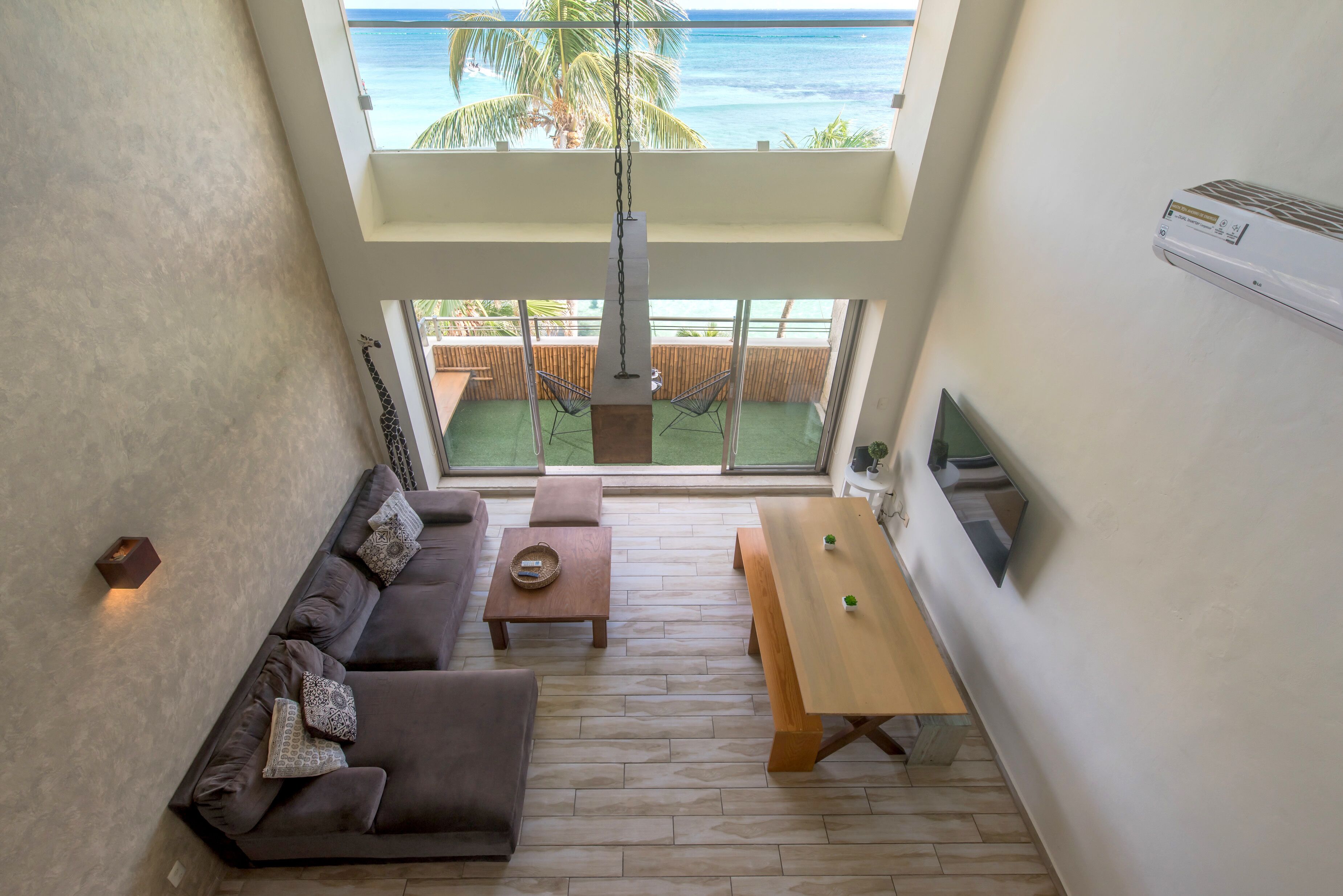 A modern living room with a sofa, coffee table, and dining table, large windows with a view of the ocean, and a hanging light fixture.