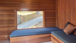 A cabin interior with a wooden sloped ceiling, a large window with a view of a rocky landscape, and a built-in bench with cushions. - 4 Acre A-Frame Private Retreat (Idyllwild)