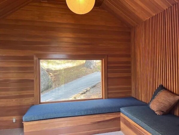 Interno di una baita con soffitto in legno inclinato, un'ampia finestra vista su un paesaggio roccioso e una panca integrata con cuscini.