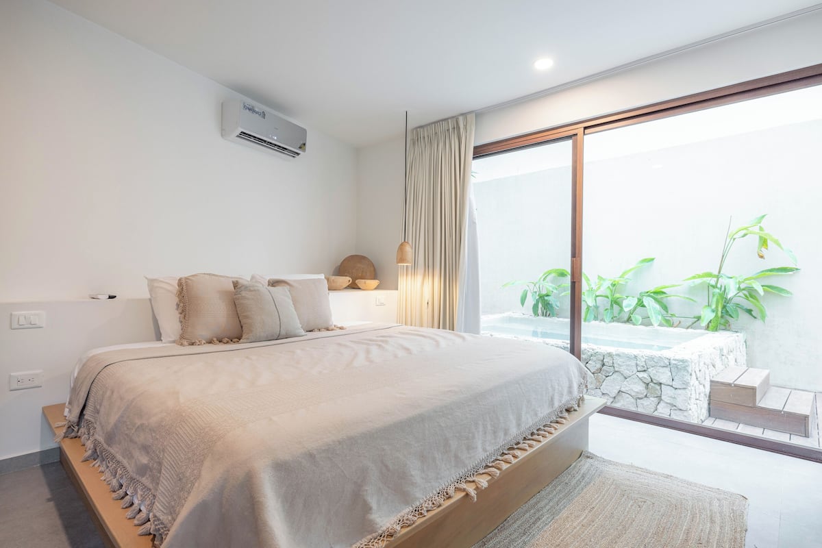 Un dormitorio moderno con una cama grande, un equipo de aire acondicionado integrado, vista a un jacuzzi y un balcón con plantas.
