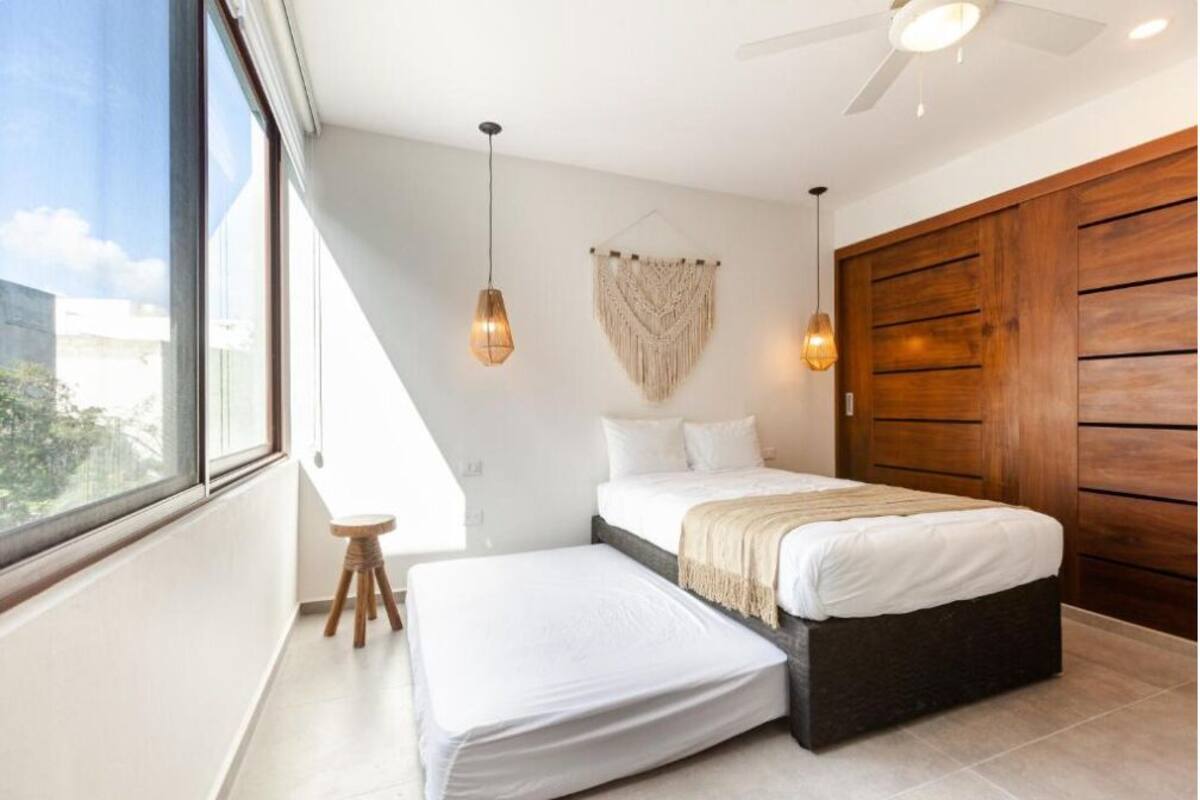 Un dormitorio con cama, un taburete pequeño, un ventilador de techo y un ventanal grande.