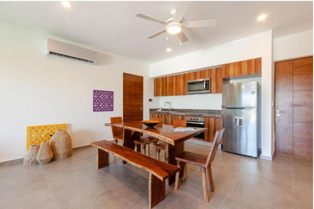 A modern kitchen with wooden cabinets, a stainless steel refrigerator, and a dining area with a wooden table and benches.